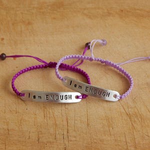 May include: Two friendship bracelets in purple and lavender with silver metal charms that read "I am ENOUGH".