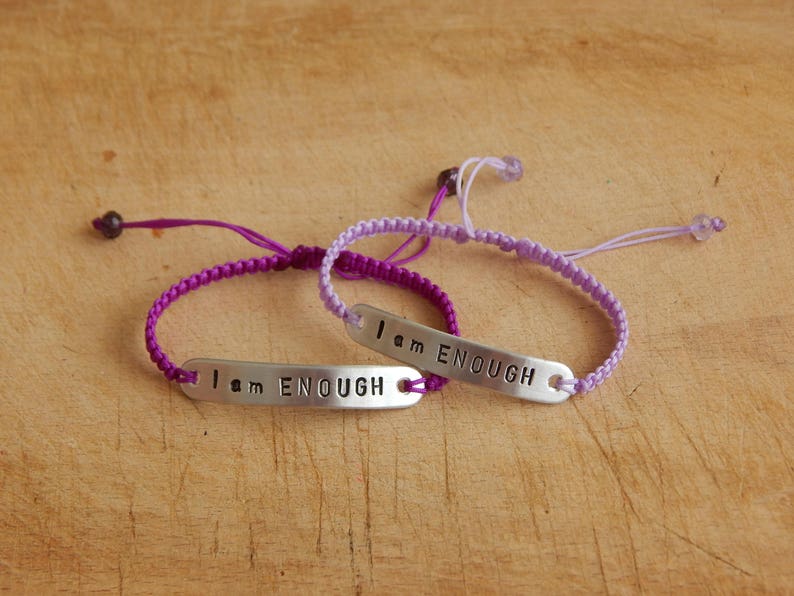 May include: Two friendship bracelets in purple and lavender with silver metal charms that read "I am ENOUGH".