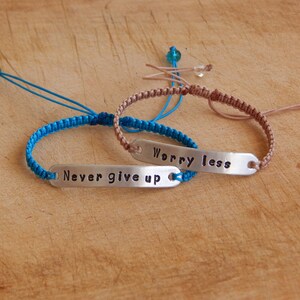 May include: Two friendship bracelets with silver charms. The blue bracelet has the text "Never give up" and the brown bracelet has the text "Worry less".