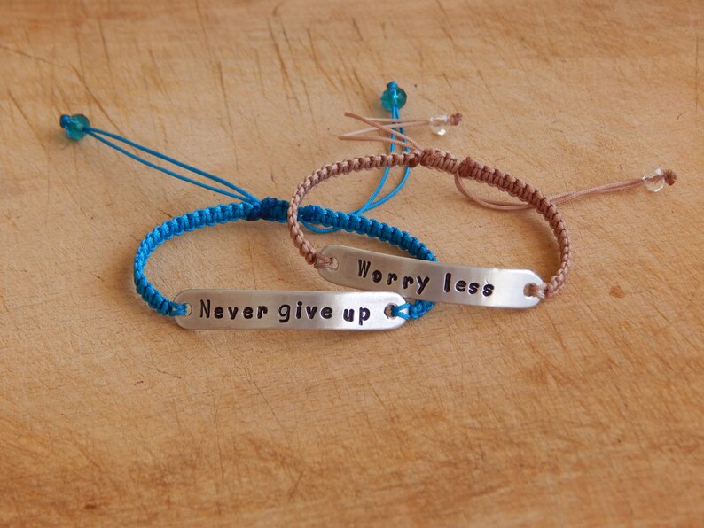 May include: Two friendship bracelets with silver charms. The blue bracelet has the text "Never give up" and the brown bracelet has the text "Worry less".