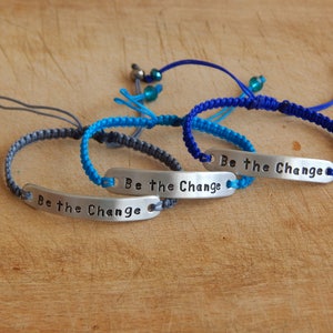 May include: Three braided bracelets with silver metal charms that say "Be the Change". The bracelets are blue, teal, and grey.