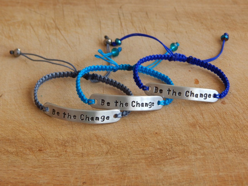 May include: Three braided bracelets with silver metal charms that say "Be the Change". The bracelets are blue, teal, and grey.