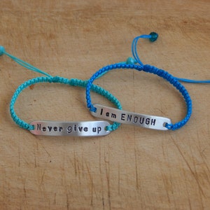 May include: Two friendship bracelets with adjustable closures. One bracelet is turquoise blue with a silver charm that reads "Never give up". The other bracelet is a darker blue with a silver charm that reads "I am ENOUGH".