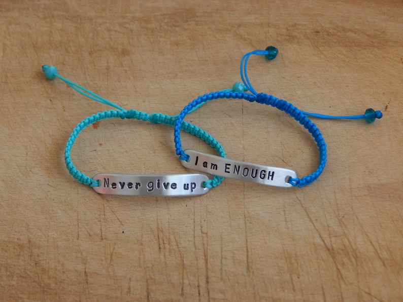 May include: Two friendship bracelets with adjustable closures. One bracelet is turquoise blue with a silver charm that reads "Never give up". The other bracelet is a darker blue with a silver charm that reads "I am ENOUGH".