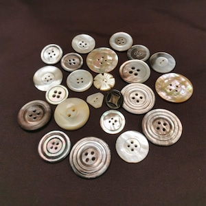 Può includere: Una collezione di bottoni di varie dimensioni e colori. I bottoni sono realizzati con materiali come madreperla e plastica, con due o quattro fori. Alcuni bottoni hanno una lucentezza perlacea, mentre altri sono marrone scuro o neri.