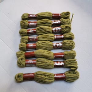 Könnte beinhalten: Eine Sammlung von olivgrünen Garnknäueln, jeweils mit einem braunen Etikett. Das Garn ist in ordentlichen Reihen auf einer weißen Oberfläche angeordnet. Die Etiketten tragen Text und ein Logo. Geeignet für Handarbeiten.