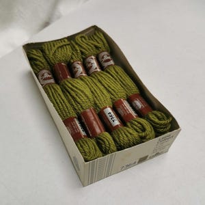 Könnte beinhalten: Eine Schachtel olivgrünes Stickgarn. Das Garn ist ordentlich in einer Pappschachtel angeordnet. Jede Spule ist mit einem braunen Etikett umwickelt. Auf der Schachtel ist die Nummer 7364 aufgedruckt.