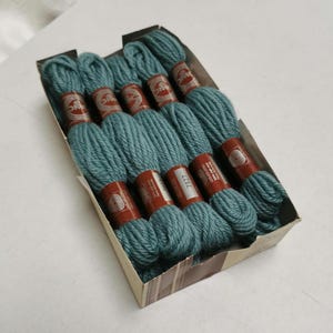 May include: A box containing teal-coloured yarn skeins. Each skein has a brown label. The yarn is neatly arranged inside the box, suggesting a craft supply or textile product.