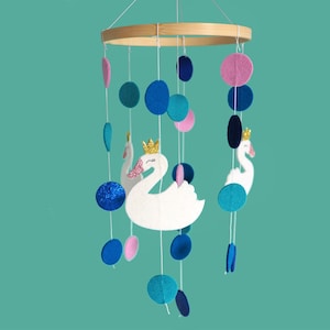 Könnte beinhalten: Ein weißer Filz-Baby-Mobile mit zwei weißen Schwänen, einer goldenen Krone und rosa und blauen Filzkreisen. Das Mobile hängt an einem Holzring.