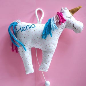 Puede incluir: Un peluche de unicornio blanco con una crin y cola rosa y azul. El unicornio tiene estrellas doradas brillantes en su cuerpo y el nombre "Henia" bordado en su costado.