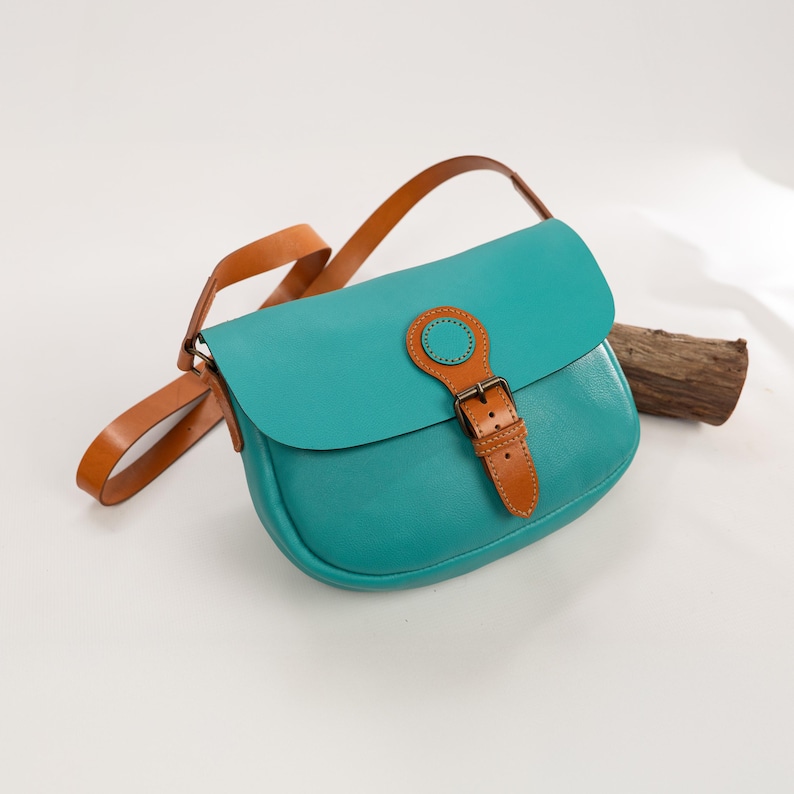May include: A teal leather crossbody bag with a brown leather strap and buckle closure. The bag has a rounded shape and a flap closure. The strap is adjustable. The bag is approximately 10 inches wide.
