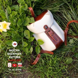 May include: A white and brown leather backpack with a buckle closure and adjustable straps sits on grass and leaves. The backpack has a flap closure and brown leather accents. The text "MOCHA BACKPACK PATTERN" is visible.