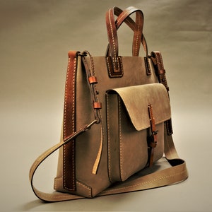 Puede incluir: Un bolso tote de cuero color topo con detalles y costuras en cuero marr&oacute;n. El bolso tiene un bolsillo frontal con solapa y una correa para el hombro. Las asas tambi&eacute;n son de cuero marr&oacute;n. El bolso tiene una forma cl&aacute;sica y estructurada.