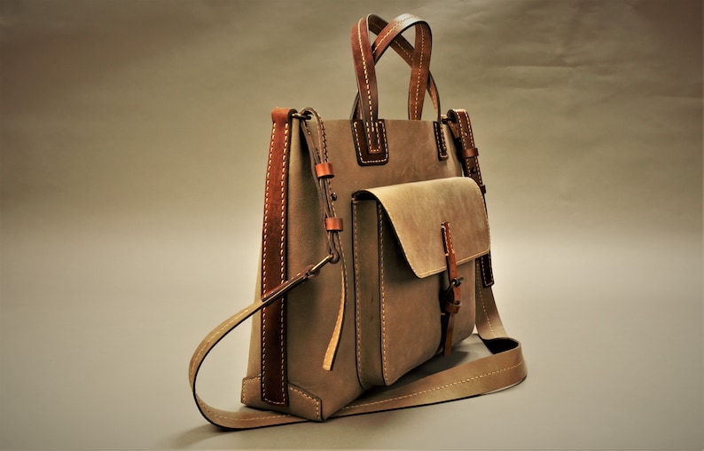 Puede incluir: Un bolso tote de cuero color topo con detalles y costuras en cuero marr&oacute;n. El bolso tiene un bolsillo frontal con solapa y una correa para el hombro. Las asas tambi&eacute;n son de cuero marr&oacute;n. El bolso tiene una forma cl&aacute;sica y estructurada.