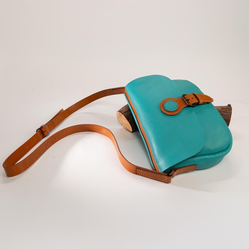 May include: A turquoise leather crossbody bag with a brown leather strap and buckle closure. The bag has a rounded flap and a contrasting brown trim. The strap is adjustable and the bag is resting on a piece of wood.