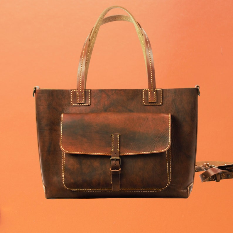 Puede incluir: Un bolso tote de cuero marr&oacute;n con un bolsillo frontal y un cierre de hebilla. El bolso tiene dos asas y una correa para el hombro. Las costuras son de color marr&oacute;n claro. El bolso est&aacute; hecho de cuero envejecido.