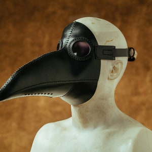 May include: A black leather plague doctor mask with a long beak and goggles. The mask is worn on a mannequin's head.