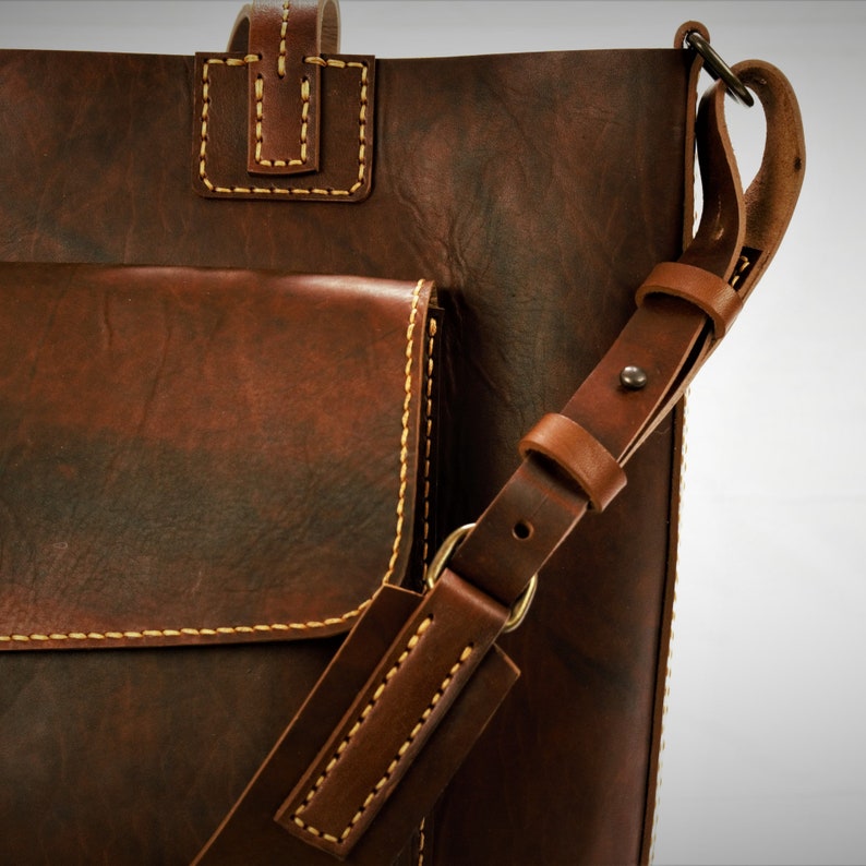 Puede incluir: Primer plano de un bolso tote de cuero marr&oacute;n con una correa para el hombro. El bolso tiene un bolsillo frontal y un asa. Costuras de color beige claro y cuero envejecido.