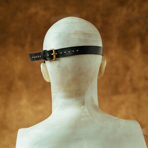 May include: Back view of a black leather headband with a gold buckle. The headband is worn on a white mannequin head.