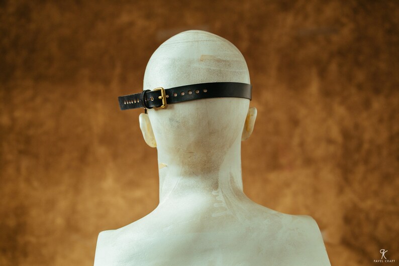 May include: Back view of a black leather headband with a gold buckle. The headband is worn on a white mannequin head.