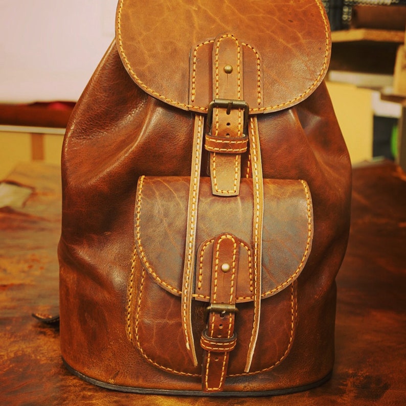 May include: A brown leather backpack with two front pockets and adjustable straps. The backpack has a flap closure with a buckle and stitching details.
