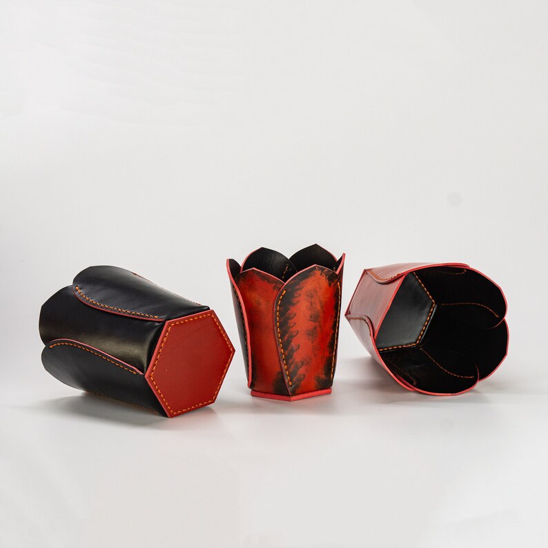 May include: Three leather pencil holders in different shapes and colors. One is black with red stitching, one is red with black stitching, and one is a tulip shape with red and black patterns.