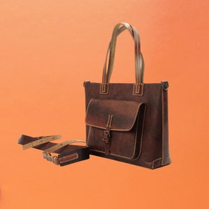Puede incluir: Un bolso tote de cuero marr&oacute;n con un bolsillo frontal y cierre de hebilla. El bolso tiene dos asas y una correa para el hombro desmontable. Las costuras son de un color m&aacute;s claro, sobre un fondo naranja.