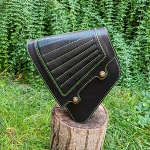 May include: Black leather motorcycle saddlebag with green stitching. The bag has a triangular shape and is secured with a metal buckle.