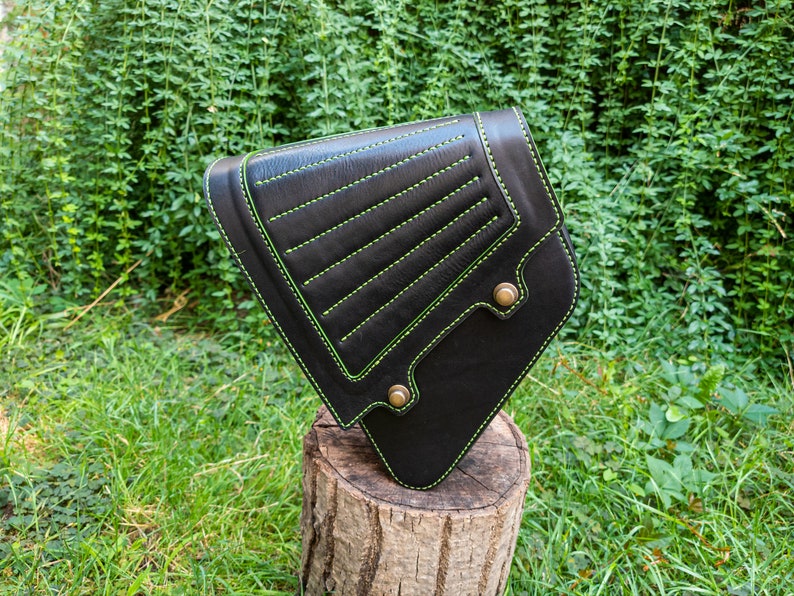 May include: Black leather motorcycle saddlebag with green stitching. The bag has a triangular shape and is secured with a metal buckle.