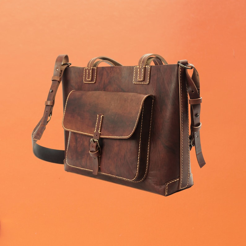 Puede incluir: Un bolso tote de cuero marr&oacute;n con un bolsillo frontal y una correa de hombro ajustable. El bolso tiene forma rectangular con un asa superior y est&aacute; cosido con hilo de color claro. El cuero tiene un aspecto envejecido.