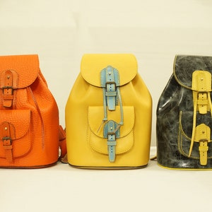 Può includere: Tre zaini in pelle di diversi colori: arancione, giallo e grigio. Ogni zaino ha una tasca frontale e una chiusura con coulisse.