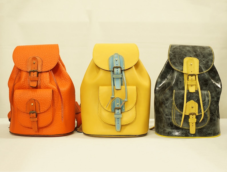 May include: Three leather backpacks in different colors: orange, yellow, and gray. Each backpack has a front pocket and a drawstring closure.
