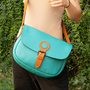 May include: A turquoise leather crossbody bag with a brown leather strap and buckle closure. The bag has a rounded shape and a flap closure. The strap is adjustable and the bag is worn over the shoulder.