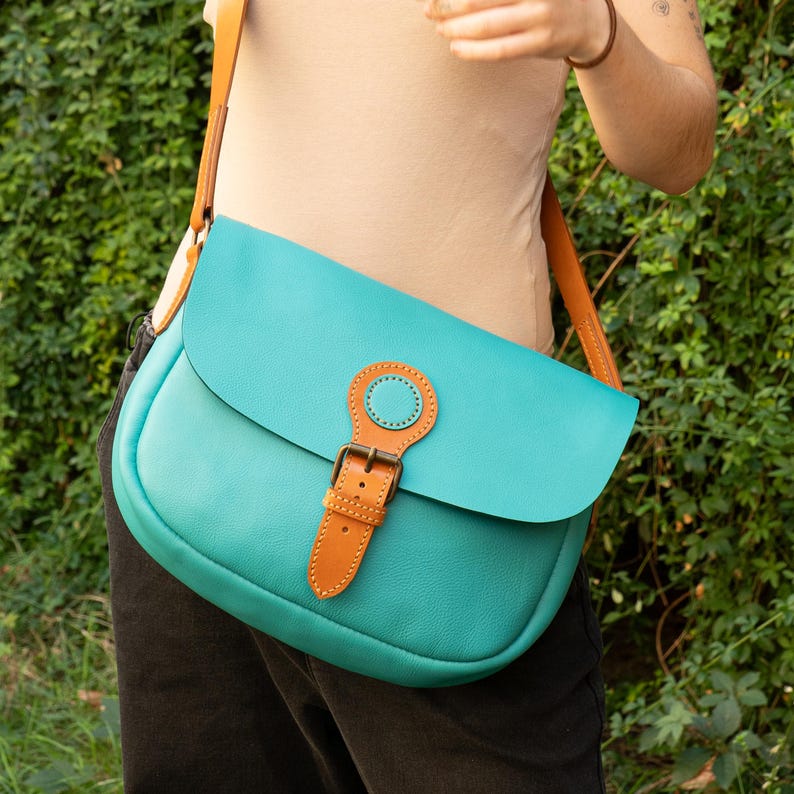 May include: A turquoise leather crossbody bag with a brown leather strap and buckle closure. The bag has a rounded shape and a flap closure. The strap is adjustable and the bag is worn over the shoulder.