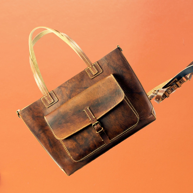 Puede incluir: Un bolso tote de cuero marr&oacute;n con un bolsillo frontal y un cierre de hebilla. El bolso tiene asas y costuras de color claro. El bolso es rectangular y parece hecho a mano. El fondo es de color naranja liso.