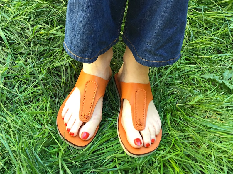 May include: Orange leather sandals with a thong-style design. The sandals have a stitched strap and a small logo. The image shows the sandals on a person's feet, with red nail polish, against a background of green grass.
