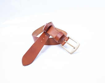 May include: A brown leather belt with a gold metal buckle. The belt is folded in half and the buckle is facing the camera.