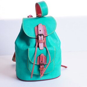 May include: A teal leather backpack with a brown leather flap and buckle closure. The backpack has yellow stitching and a small pocket on the front.