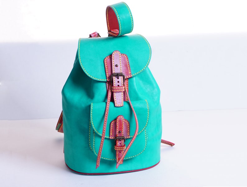 May include: A teal leather backpack with a brown leather flap and buckle closure. The backpack has yellow stitching and a small pocket on the front.
