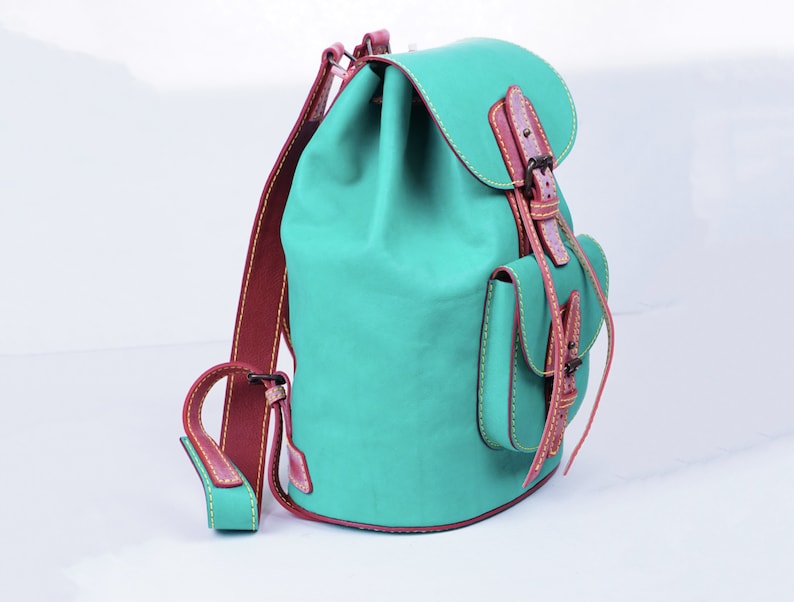 May include: A teal blue leather backpack with brown leather straps and a buckle closure. The backpack has a small front pocket with a flap and a drawstring closure.