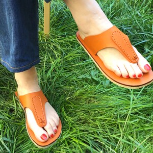 May include: A pair of orange leather sandals with a thong-style design. The sandals have a light-colored sole and are worn on feet with red nail polish, set against a backdrop of green grass.