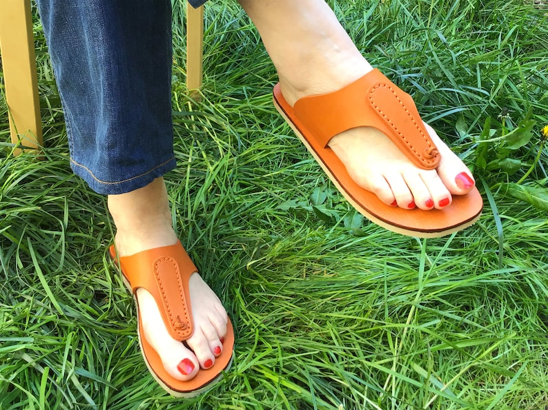 May include: A pair of orange leather sandals with a thong-style design. The sandals have a light-colored sole and are worn on feet with red nail polish, set against a backdrop of green grass.