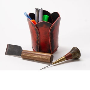 May include: A red leather pen holder with a flower shape, filled with pens and markers.  A small wooden tool with a metal tip and a brown wooden handle is also pictured.