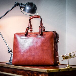 May include: A brown leather briefcase with a top handle and a zipped closure. The briefcase is sitting on a wooden table with brass trim.