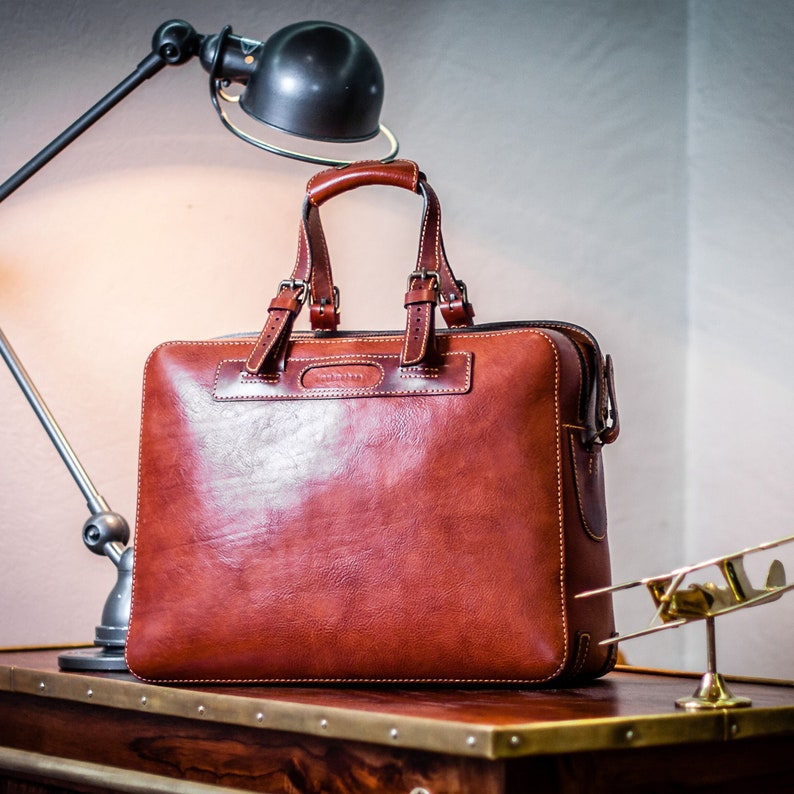 May include: A brown leather briefcase with a top handle and a zipped closure. The briefcase is sitting on a wooden table with brass trim.