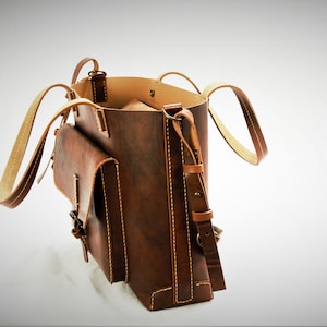 Puede incluir: Un bolso tote de cuero marr&oacute;n con un bolsillo con solapa y una correa de hombro ajustable. El bolso tiene costuras y asas de color claro. El interior es de un tono marr&oacute;n m&aacute;s claro. El bolso est&aacute; abierto, revelando el interior.
