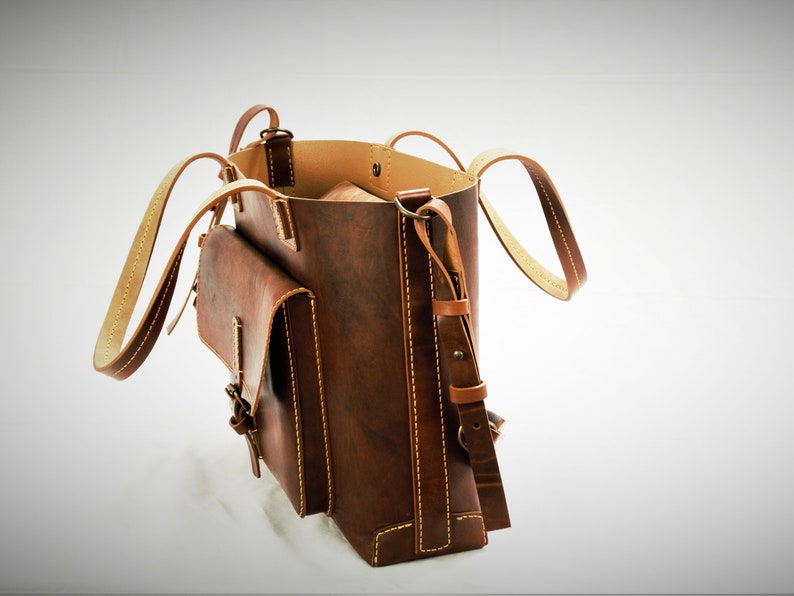 Puede incluir: Un bolso tote de cuero marr&oacute;n con un bolsillo con solapa y una correa de hombro ajustable. El bolso tiene costuras y asas de color claro. El interior es de un tono marr&oacute;n m&aacute;s claro. El bolso est&aacute; abierto, revelando el interior.