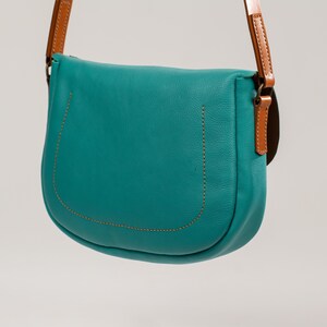 May include: A teal-colored leather crossbody bag with a curved design. The bag features a brown leather strap and accents, with visible stitching. The bag is designed with a flap closure and a rounded bottom.