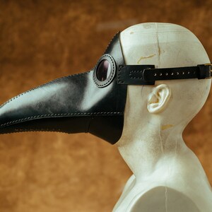 May include: A black leather plague doctor mask with a long beak and a round eye piece. The mask is worn on a mannequin head.