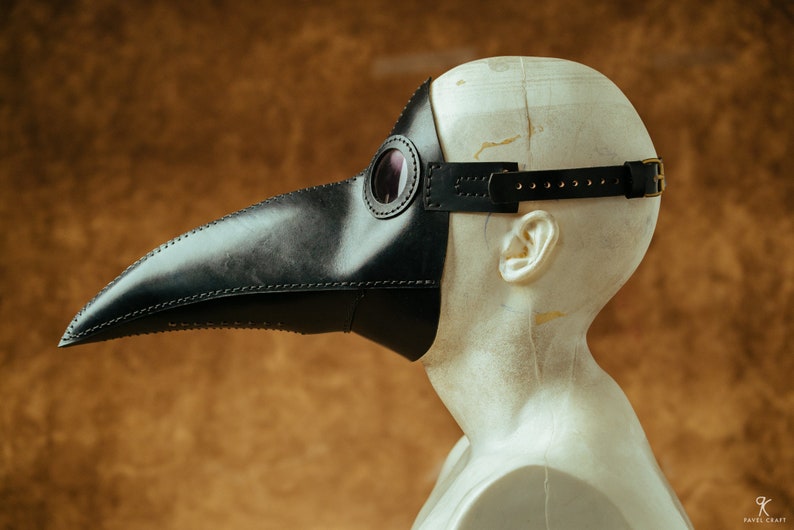 May include: A black leather plague doctor mask with a long beak and a round eye piece. The mask is worn on a mannequin head.